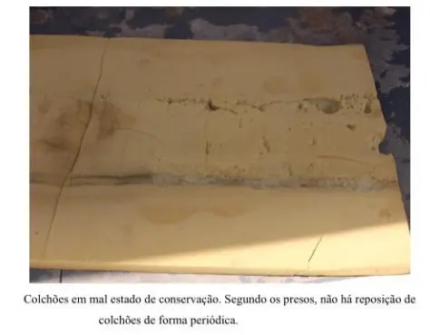 Foto de um colchão de espuma amarelo, bastante gasto e rasgado ao meio, com partes afundadas e rasuras profundas. O colchão está apoiado no chão de cimento, sob luz natural.