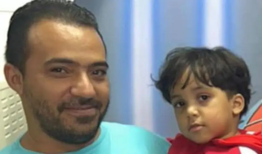 Foto colorida mostra um homem adulto segurando uma criança pequena no colo. O homem tem pele morena, cabelo curto e barba, e veste camiseta azul-clara. A criança, também de pele morena, tem cabelo curto e escuro e usa uma blusa vermelha. Ambos olham para a câmera. O fundo é um ambiente interno, com parede clara e cortina azul.