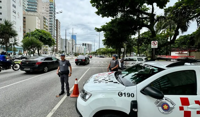 Foto colorida mostra uma avenida larga, com prédios altos ao fundo e árvores nas calçadas. Em primeiro plano, uma viatura da Polícia Militar de São Paulo está parada na via, com o logotipo da corporação visível. Três policiais militares uniformizados aparecem na cena, posicionados próximos à viatura e a cones de trânsito, enquanto veículos circulam pela avenida. O dia está nublado.