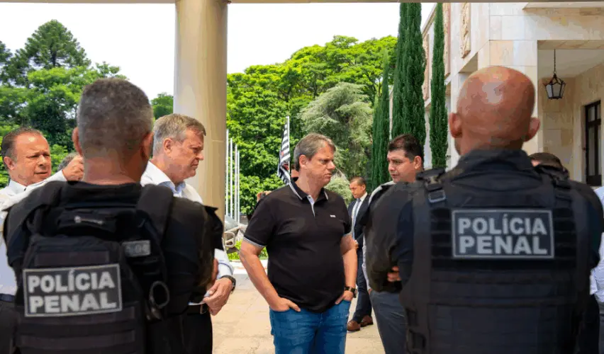 Imagem mostra o governador de São Paulo em área externa de prédio oficial, cercado por homens de terno e por agentes com coletes escritos “Polícia Penal” nas costas, em clima de conversa, com árvores e bandeira ao fundo.