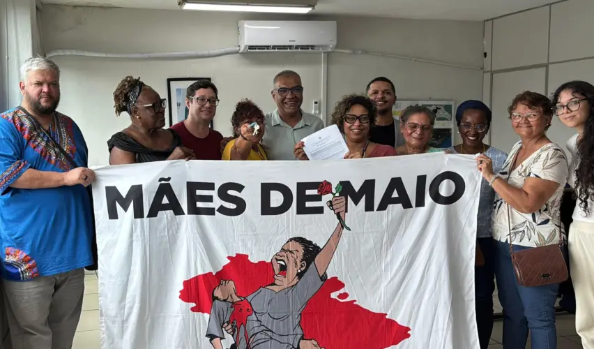 Foto em ambiente interno mostra um grupo de cerca de dez pessoas em pé, lado a lado, segurando uma faixa branca com a inscrição “Mães de Maio” e a ilustração de uma mulher erguendo uma flor. Algumas pessoas sorriem e uma delas segura um documento. O grupo participa de uma articulação institucional ligada ao projeto do Centro de Memória da Violência de Estado, em Santos (SP).