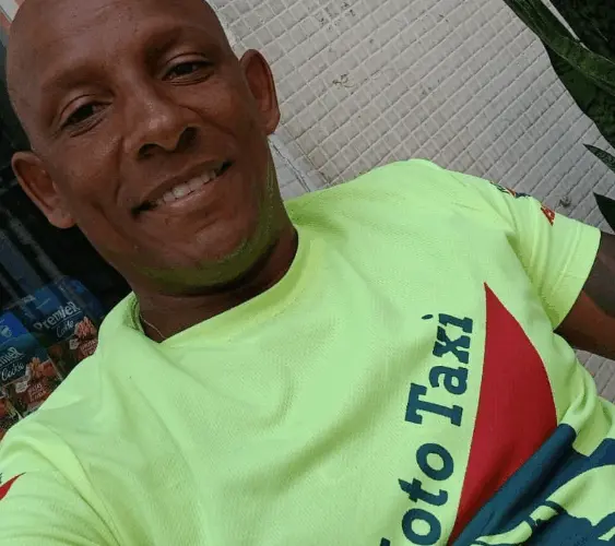 Homem negro, de meia-idade, com a cabeça raspada, sorri levemente para a câmera. Ele veste uma camiseta verde fluorescente com estampa em vermelho e azul. A foto é um retrato feito de perto, em ambiente externo, com uma parede clara ao fundo.