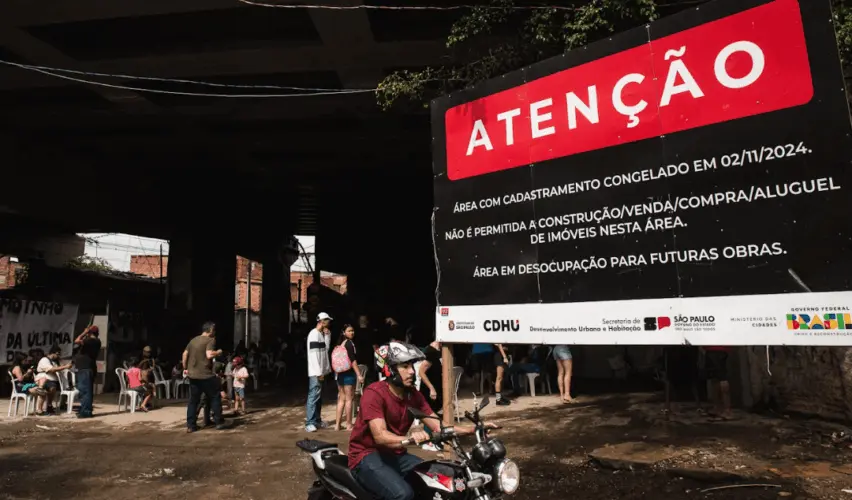 Foto de uma das entradas da favela do Moinho, em São Paulo, com grande placa preta e vermelha escrito “ATENÇÃO” anunciando congelamento de cadastro e desocupação para futuras obras, enquanto moradores circulam e um homem passa de moto em frente à placa, sob o viaduto.