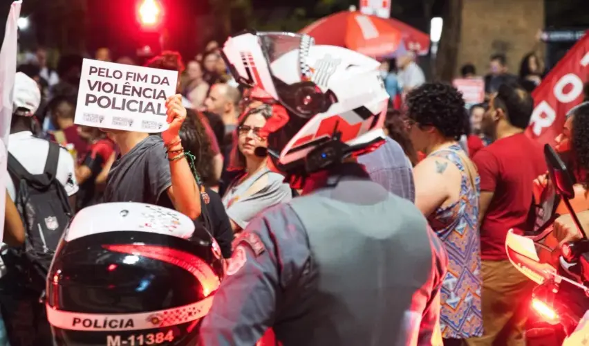 Em uma manifestação noturna em São Paulo, pessoas se concentram em via pública enquanto policiais militares, usando capacetes e motos, acompanham o ato. Em primeiro plano, um manifestante segura um cartaz com a frase “Pelo fim da violência policial”. Luzes vermelhas de viaturas iluminam a cena, e há outras pessoas ao fundo observando ou participando do protesto.