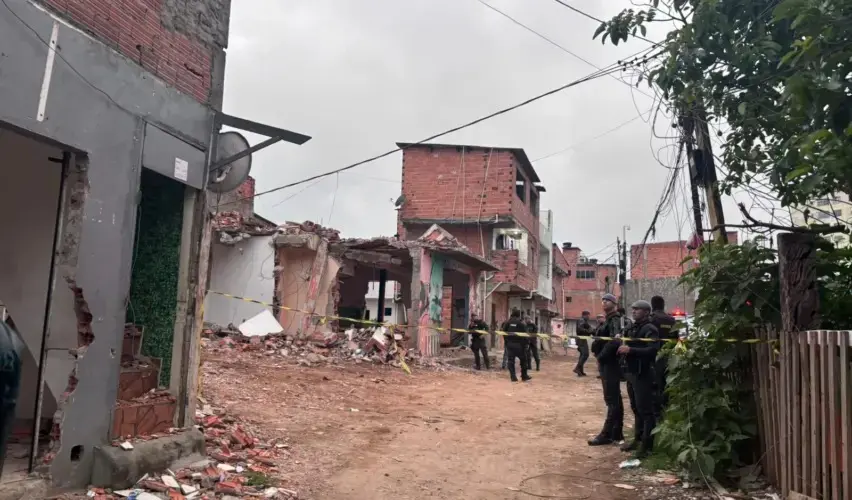 Vista de uma área da favela do Moinho, no centro de São Paulo, com imóveis parcialmente demolidos e entulho espalhado pelo chão. Uma faixa de isolamento delimita o local, onde policiais militares permanecem posicionados ao longo da via de terra. Ao fundo, aparecem casas de alvenaria e fiação aérea, sob céu nublado.